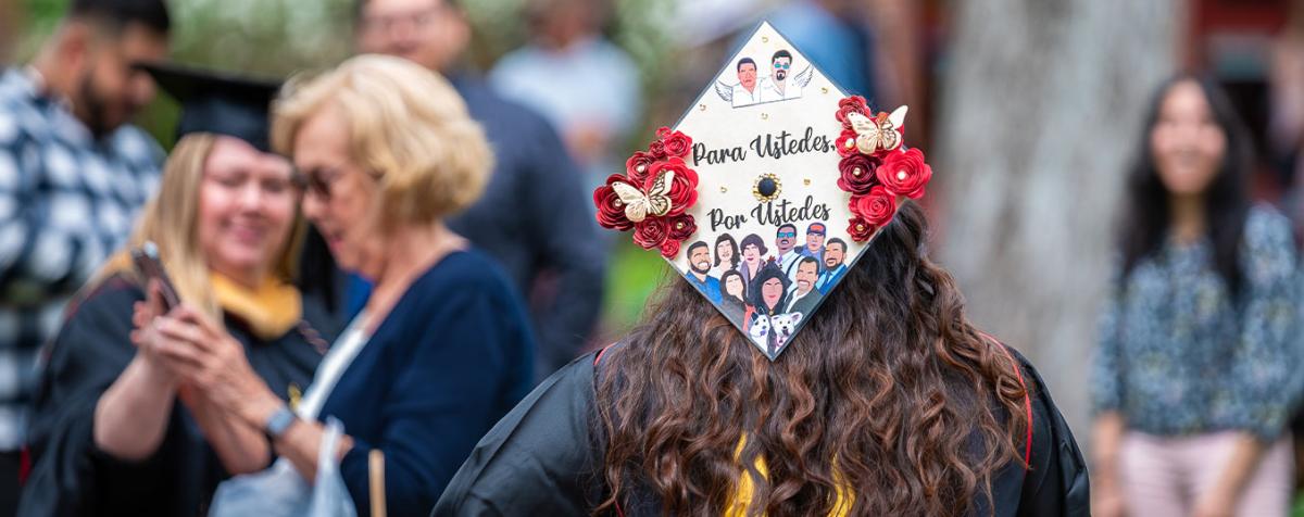 Student in regalia with a cap that reads "Para Ustedes, Por Ustedes" (For You, Because of You) with illustrations of friends and family